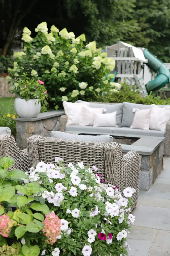 Outdoor patio with wicker furniture, cushioned seating, a fireplace, and blooming flowers. A playground with a slide is visible in the background surrounded by greenery.