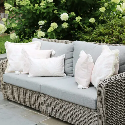 A wicker outdoor sofa with gray cushions and several white patterned throw pillows sits on a patio surrounded by green plants and flowers.