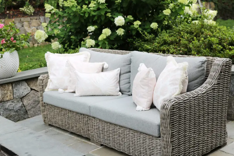 A wicker outdoor sofa with gray cushions and several white patterned throw pillows sits on a patio surrounded by green plants and flowers.