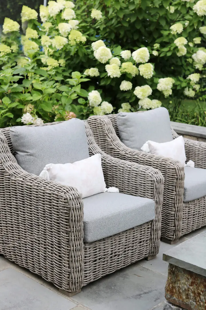 Two wicker patio chairs with gray cushions and white pillows sit on a stone patio, surrounded by green plants with white flowers.