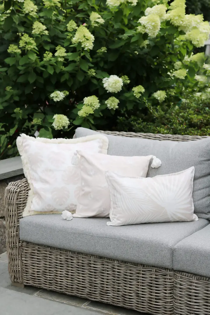 Outdoor wicker sofa with gray cushions and three decorative white pillows. Green foliage with blooming white flowers in the background.