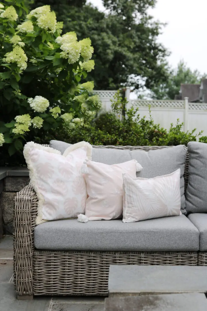 A wicker outdoor couch with light gray cushions adorned with white and pastel pink decorative pillows sits on a patio. Green hydrangea bushes and a wooden fence are visible in the background.