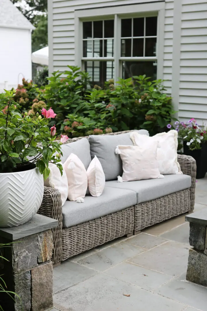 A wicker outdoor sofa with gray cushions and white patterned pillows sits on a stone patio. A large white plant pot with pink flowers is nearby. Shrubs and a window are in the background.