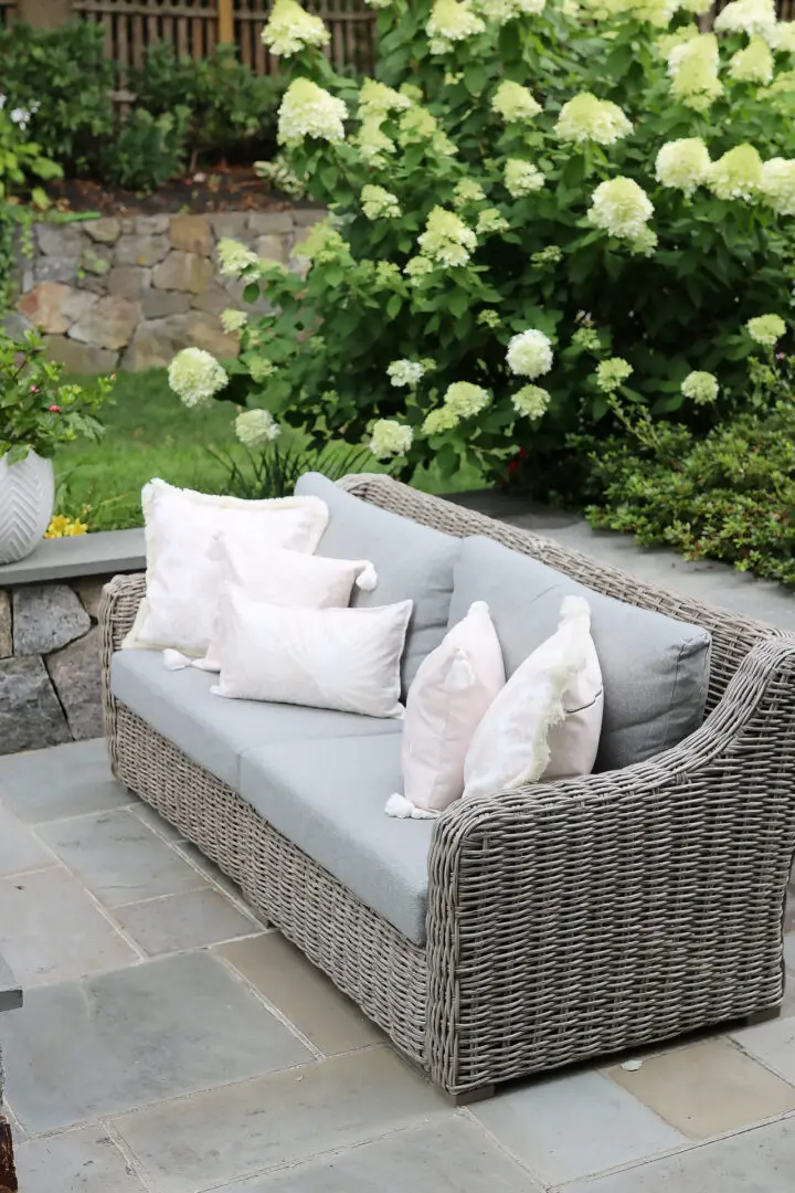 A gray outdoor wicker sofa with light-colored throw pillows is placed on a stone patio, surrounded by greenery and blooming hydrangeas.