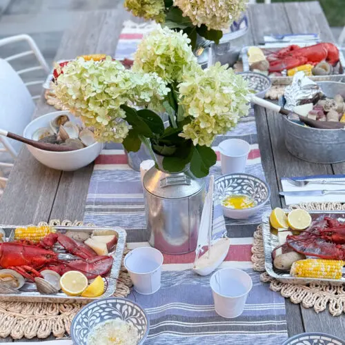 A dining table is set outdoors with seafood platters, including lobsters, corn, and clams. White hydrangea centerpieces decorate the table, and metal buckets with additional food items are present.