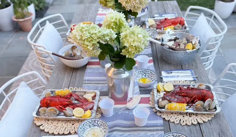 A dining table is set outdoors with seafood platters, including lobsters, corn, and clams. White hydrangea centerpieces decorate the table, and metal buckets with additional food items are present.