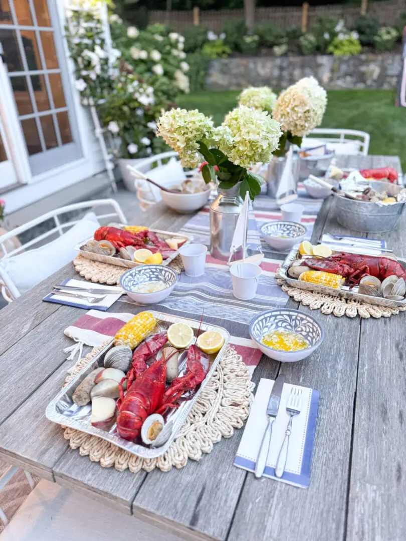 Outdoor dining table set with seafood plates featuring lobsters, clams, corn, and potatoes, along with bowls of butter, silverware, and floral centerpieces.