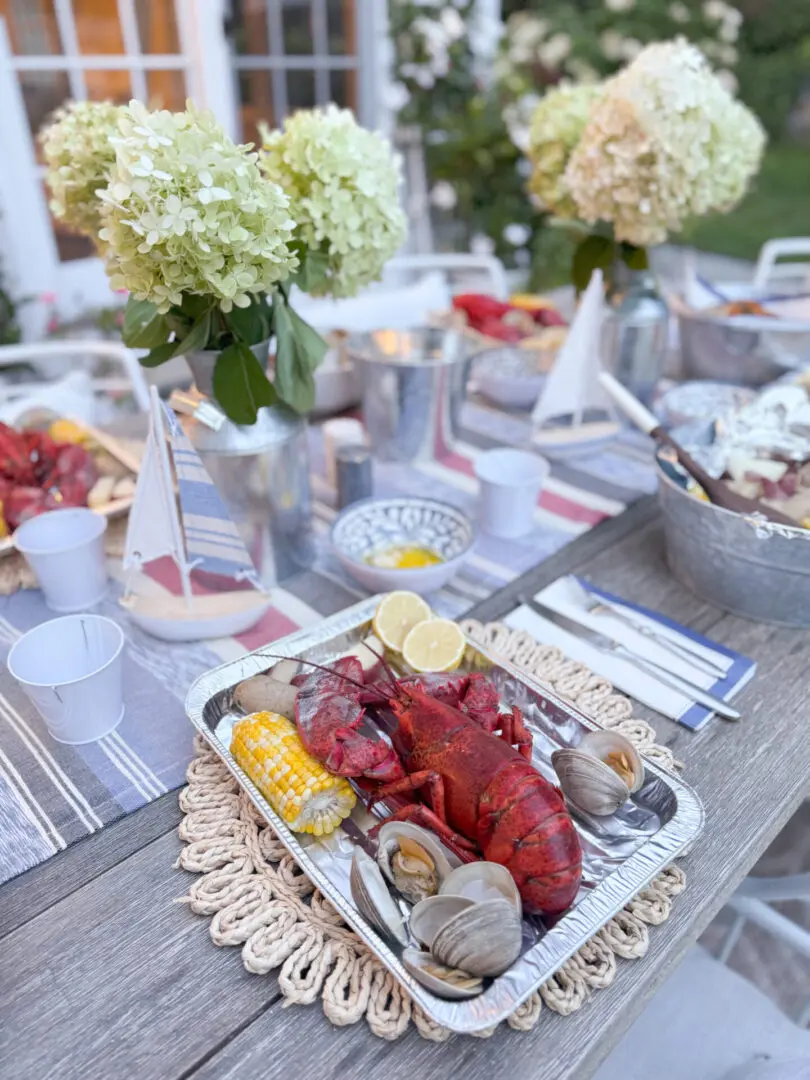 Savoring summer with an end-of-summer lobster boil night and to reflect on the coming up school year and to set goals and new ambitions. Darling Darleen Top CT Lifestyle Blogger A seafood meal including a whole lobster, clams, and corn on the cob is served on a table decorated with flowers and miniature sailboats.