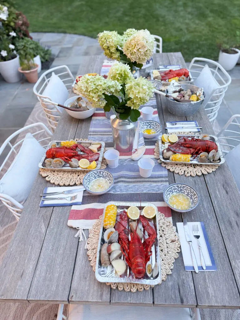 Savoring summer with an end-of-summer lobster boil night and to reflect on the coming up school year and to set goals and new ambitions. Darling Darleen Top CT Lifestyle Blogger Outdoor dining table set with plates of lobster, clams, corn on the cob, and lemon wedges, a centerpiece of white flowers, and place settings with napkins and utensils on a wooden table.