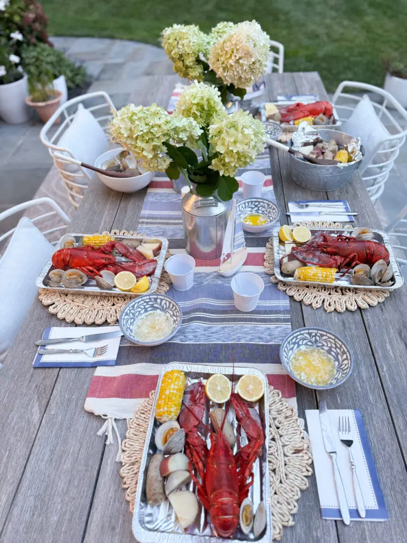 Savoring summer with an end-of-summer lobster boil night and to reflect on the coming up school year and to set goals and new ambitions. A wooden table set with lobster dishes, corn, clams, lemon slices, flowers in vases, and dining utensils. Four place settings are arranged for a meal outdoors.