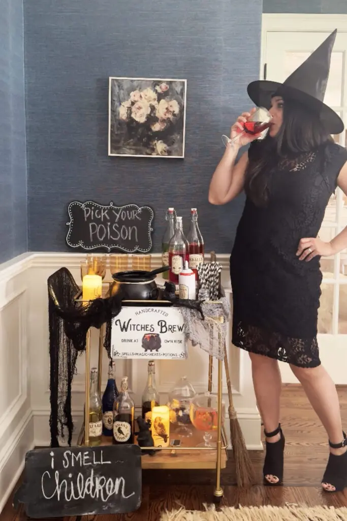 Transform Your Home into a Witch’s Haven! Woman in a witch hat drinks from a glass near a Halloween-themed drink station with bottles, signs, and decorations.