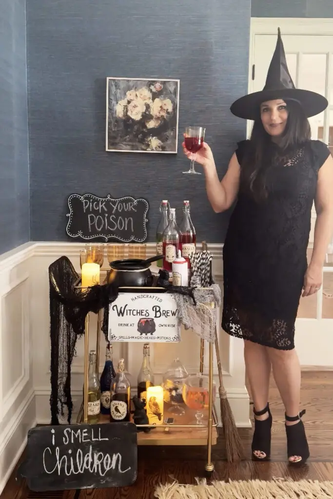 Transform Your Home into a Witch’s Haven! A woman in a witch hat stands by a Halloween-themed drink cart. She holds a glass of red wine. There are potions, candles, and a “Pick Your Poison” sign on the cart.