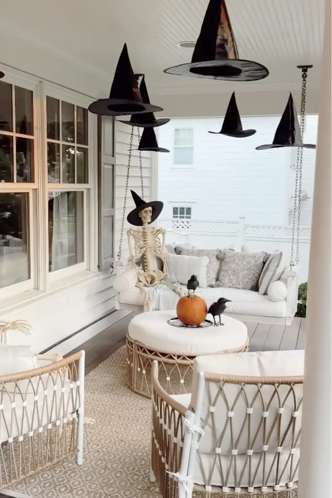 Transform Your Home into a Witch’s Haven! A porch with a white sofa, skeleton, and pumpkin centerpiece. Witch hats hang from the ceiling, and two chairs face the table with black crows on the pumpkin.