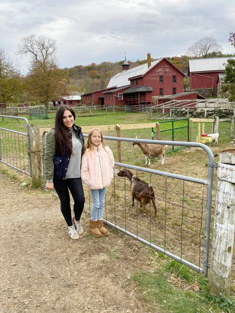 Your Guide to the Best Fall Activities Near Fairfield County, Connecticut in the month of October from pumpkin patches to haunted houses! A woman and child stand by a metal gate with goats in a rural farm setting. Red barns and trees are in the background.