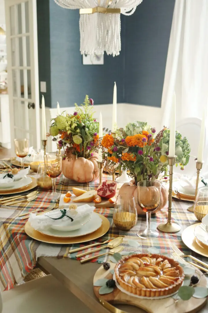 A dining table with a fall-themed setup, featuring plates, glasses, flower arrangements, pumpkins, candles, and an apple tart. The table is covered with a plaid tablecloth.