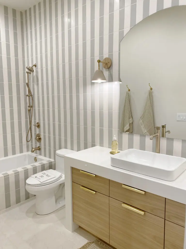 2025 Bathroom Trends Modern bathroom with vertical striped tiles, wall-mounted light, and a large mirror. Features a wooden vanity with a white vessel sink and faucet, toilet, and shower-tub with brass fixtures.