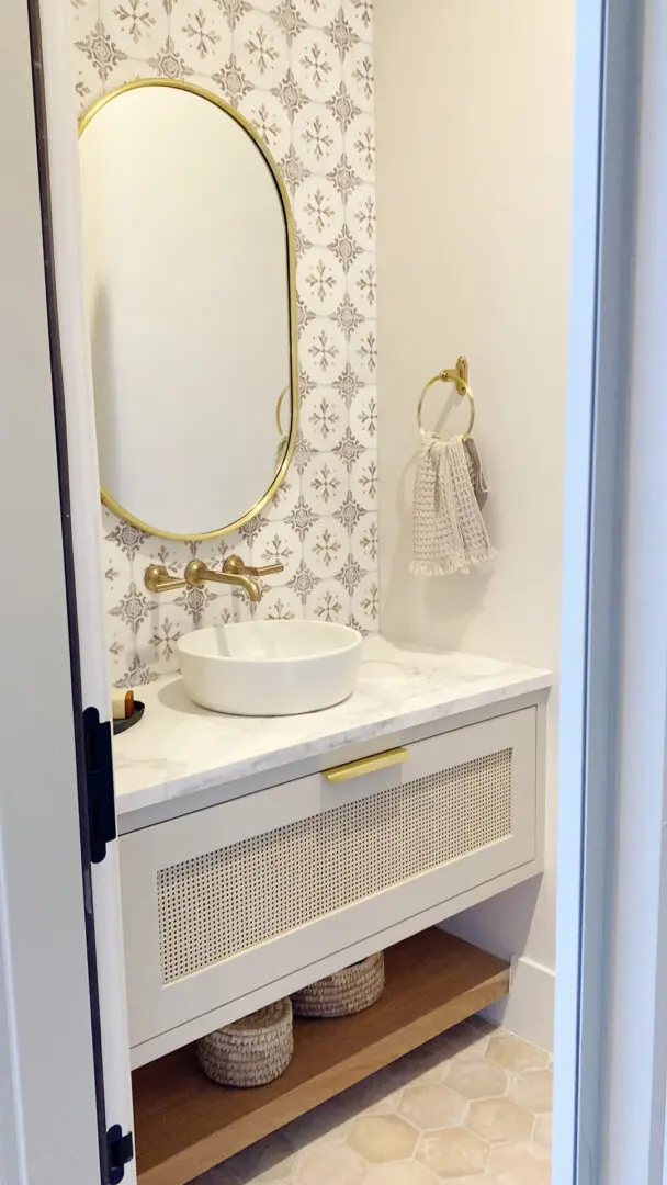 2025 Bathroom Trends Small bathroom with a white vanity, round basin, and gold fixtures. Oval mirror and patterned tile backsplash, with a decorative hand towel on a ring next to the sink.