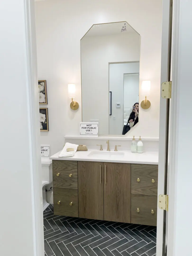 2025 Bathroom Trends A modern bathroom with a wooden vanity, two gold faucets, hexagonal mirror, wall sconces, and black herringbone floor. A small sign on the counter reads "Not for Public Use.