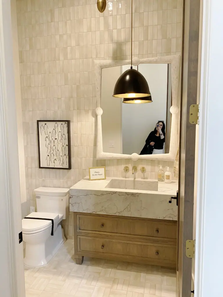 2025 Bathroom Trends Bathroom with tiled walls, a wooden vanity with a marble countertop, a toilet, a wall mirror, and a black pendant light. A person is visible in the mirror taking a photo.