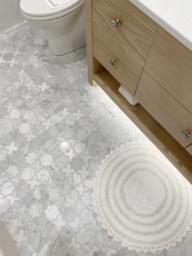 2025 Bathroom Trends Bathroom with white and gray patterned tile floor, wooden vanity with drawers, a round white rug, and a toilet in the background.