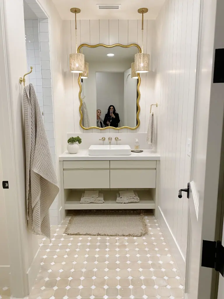 2025 Bathroom Trends Bathroom with cream and white decor, featuring a gold-framed mirror, white vanity, and pendant lights. Beige patterned floor and towels. Two people reflected in the mirror.