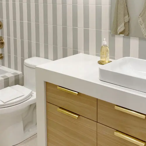 Bathroom with striped tile walls, a bathtub, toilet, and modern sink on a wooden vanity. There's a gold faucet and soap dispenser, and towels hang near the mirror.