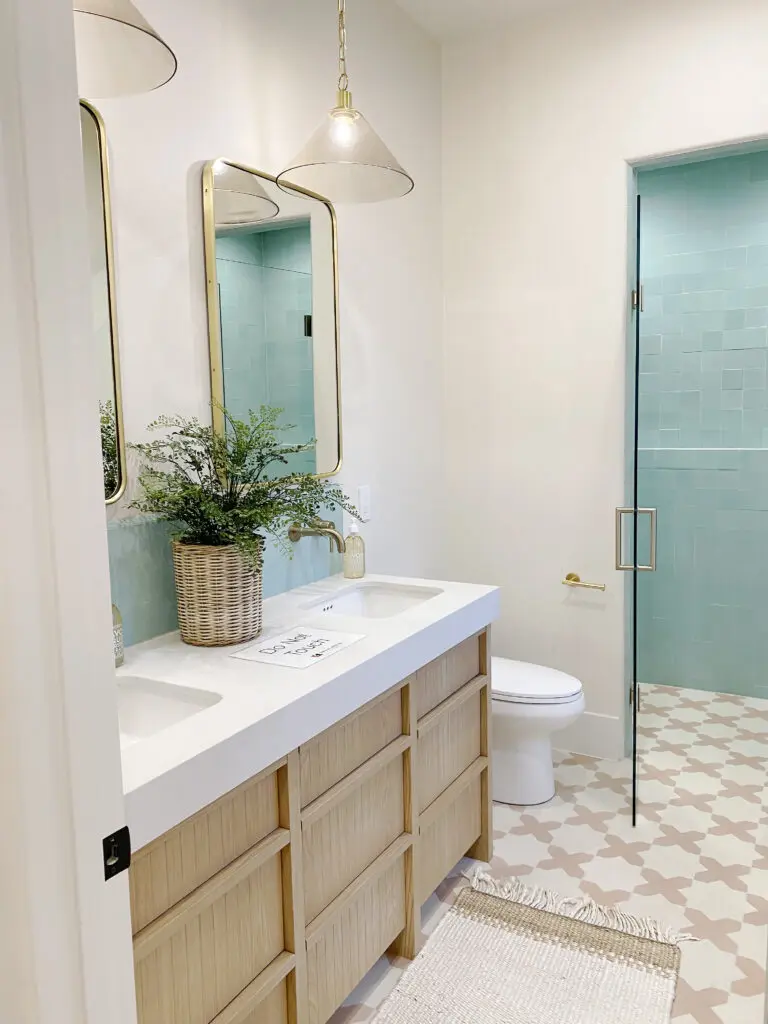 2025 Bathroom Trends Modern bathroom with double sinks, rectangular mirrors, a green plant on the counter, pendant lights, and a glass shower with blue tiles. Beige patterned floor.