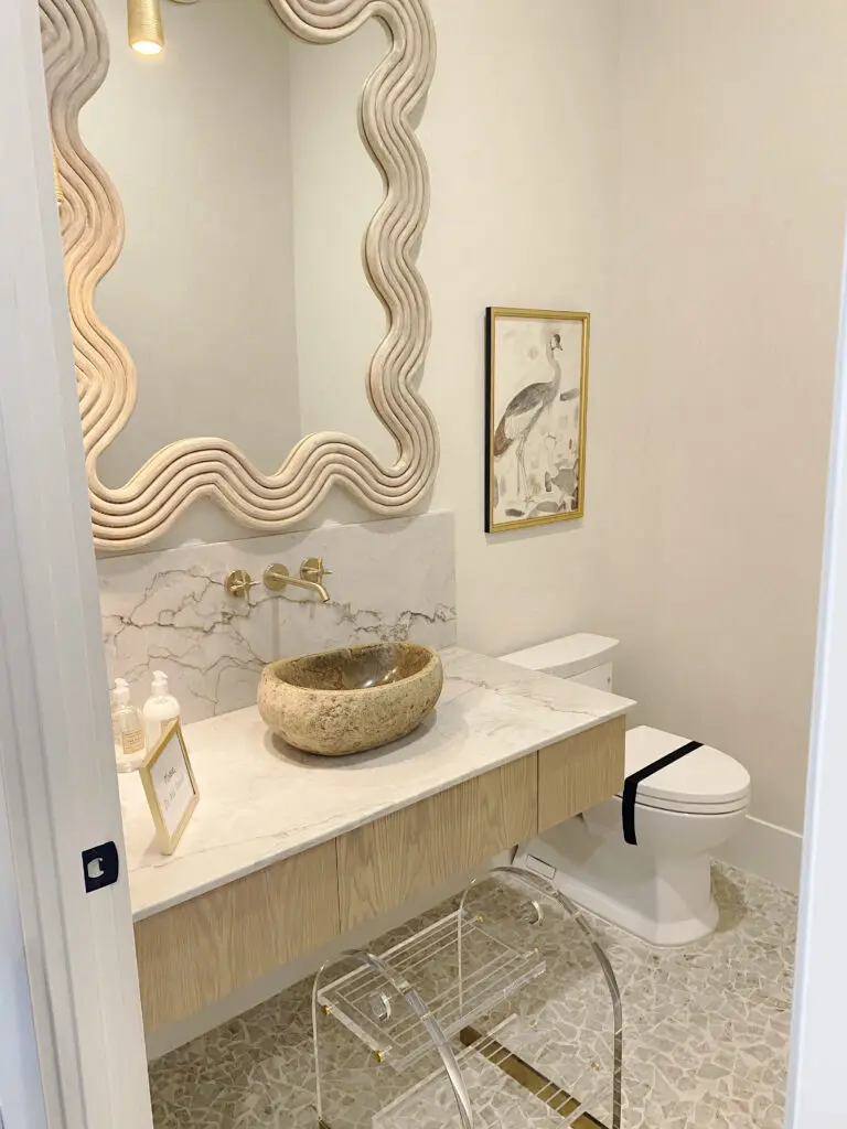 2025 Bathroom Trends Small, modern bathroom with a stone sink on a marble countertop, ornate mirror, wall-mounted faucet, framed bird picture, and a transparent chair next to a toilet.