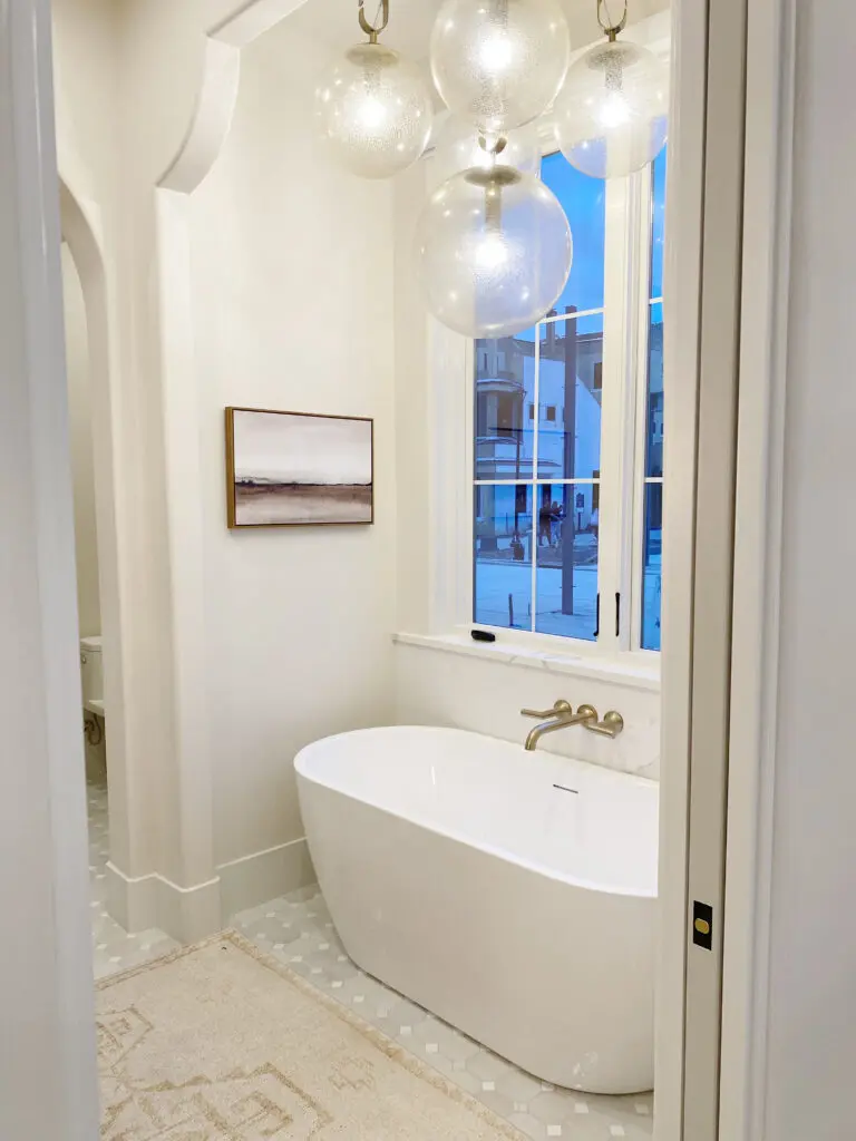 2025 Bathroom Trends Bathroom with a freestanding white tub, wall-mounted artwork, and a cluster of spherical pendant lights. Large window with view of buildings outside. Beige floor with patterned tiles.