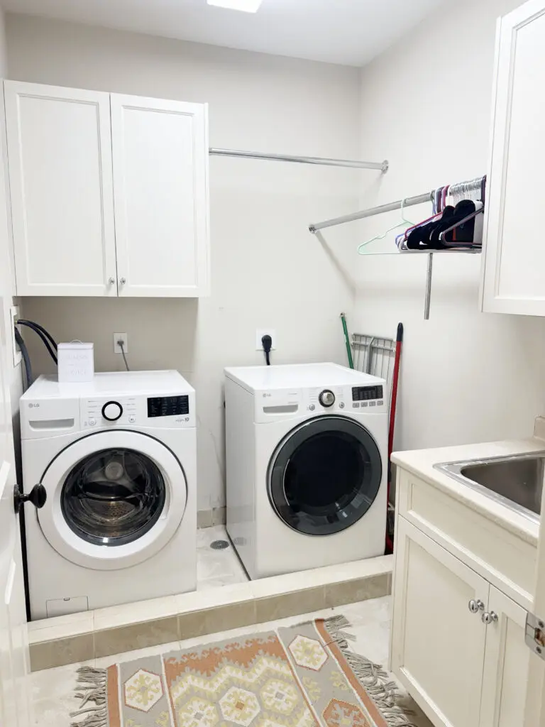 Laundry room with a white washer and dryer, cabinets, a sink, clothes rack, and cleaning tools. A patterned rug is on the floor.