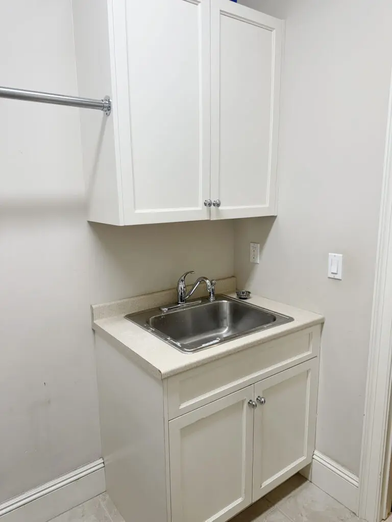 A laundry room with white cabinets, stainless steel sink, and countertop. A silver rod is mounted on the wall above the cabinets.