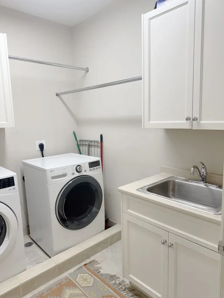 A laundry room with a washing machine, dryer, and a countertop with a sink. White cabinets are mounted above the sink and appliances. A hanging rod is installed on the wall.