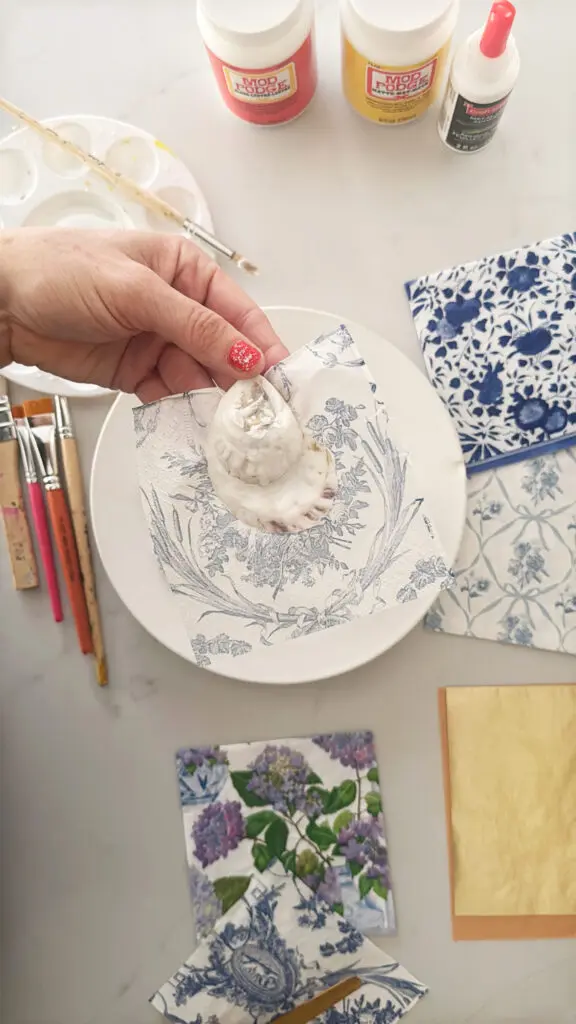 A hand holding a seashell covered in a floral napkin, surrounded by brushes, Mod Podge containers, and patterned napkins on a table.