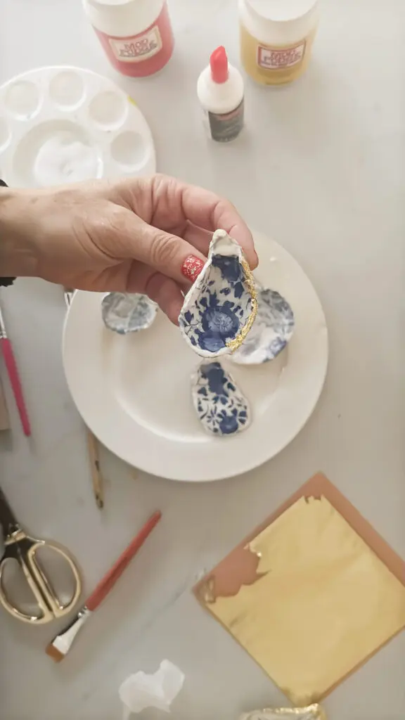 A hand holds a decorative oyster shell with blue patterns and gold edging. Other similar shells, art supplies, and gold leaf squares are on a table.