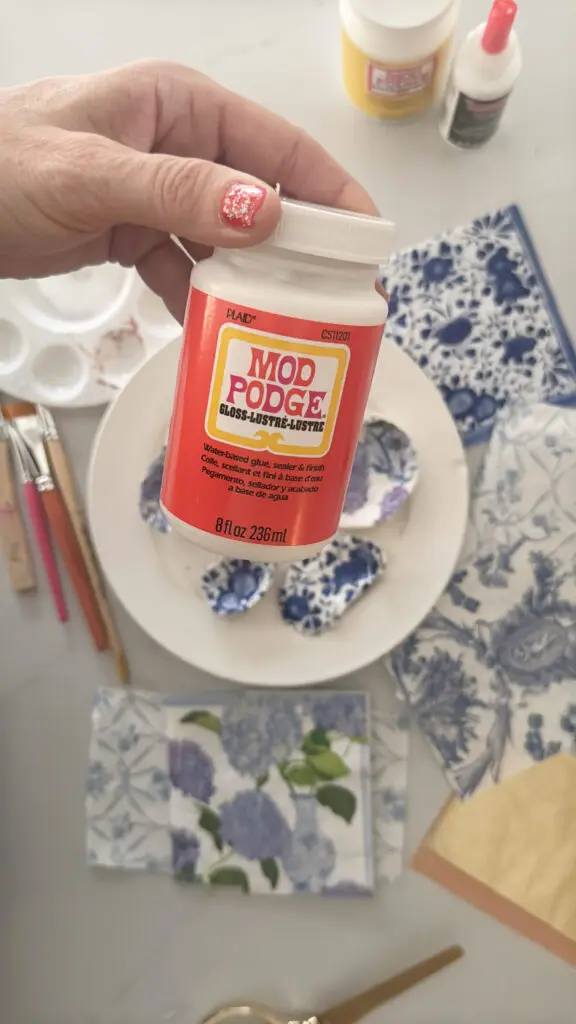 Hand holding a container of Mod Podge above a table with decorative papers, paintbrushes, and oyster shells.