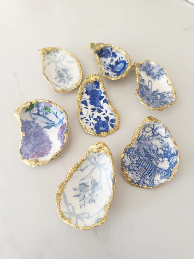 Seven decorative oyster shells with blue and white floral patterns and gold edges are arranged on a white surface.