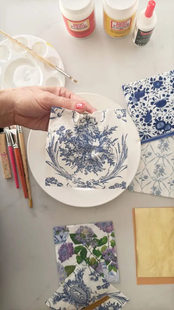 A hand holds a decorative blue and white napkin over a plate. Art supplies, including glue, paint, and brushes, are scattered around.