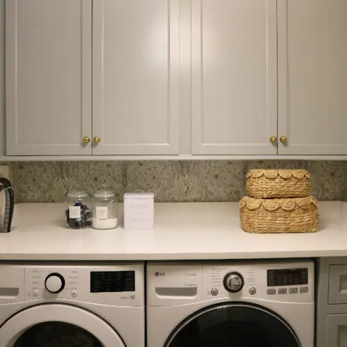Modern laundry room with front-loading washer and dryer, gray cabinets, woven baskets, laundry supplies, and patterned wallpaper.