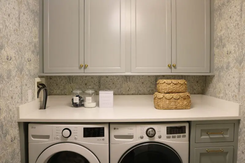 Modern laundry room with front-loading washer and dryer, gray cabinets, woven baskets, laundry supplies, and patterned wallpaper.