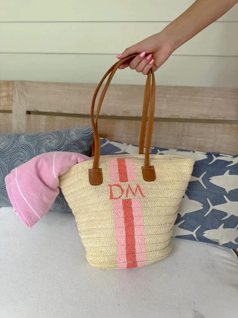 A hand holding a woven straw tote bag with brown handles, pink stripes, and the initials "DM," resting on a couch with patterned pillows and a pink towel. || Easy Paint Craft Project to Personalize Your Summer Bag with a DIY hand-painted monogram straw bag that is just so cute!