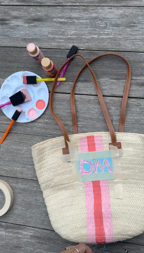 A straw tote bag with brown handles is being painted with pink stripes and blue letters "DM" using paints, brushes, and painter's tape on a wooden surface.
