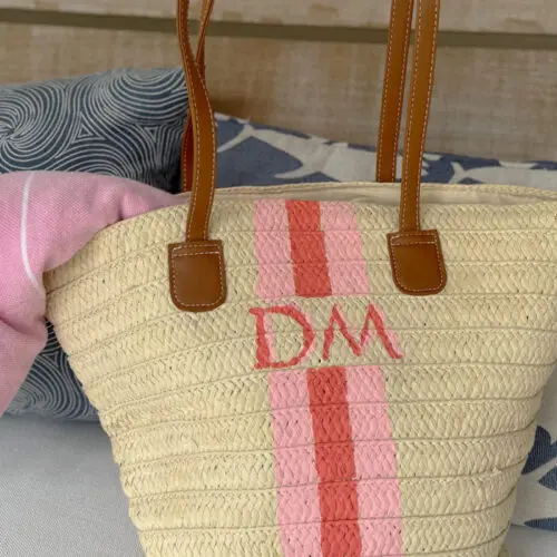 A straw tote bag with brown handles, pink stripes, and the initials "DM" rests on a couch beside blue and white fish-patterned and swirled pillows, with a pink cloth inside.