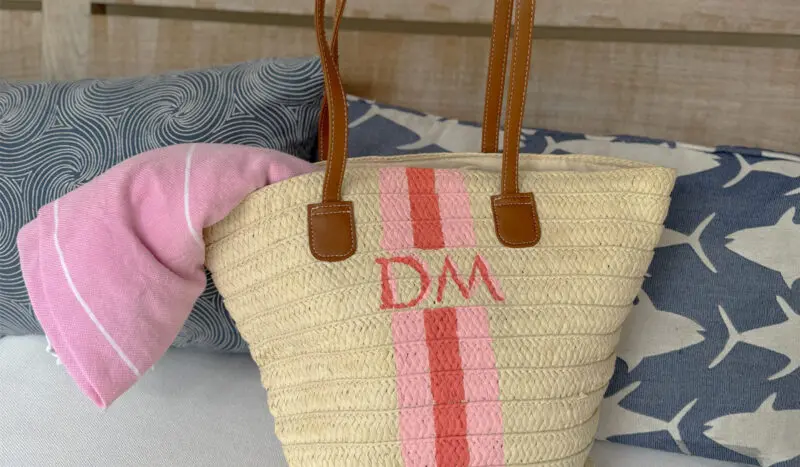 A straw tote bag with brown handles, pink stripes, and the initials "DM" rests on a couch beside blue and white fish-patterned and swirled pillows, with a pink cloth inside.