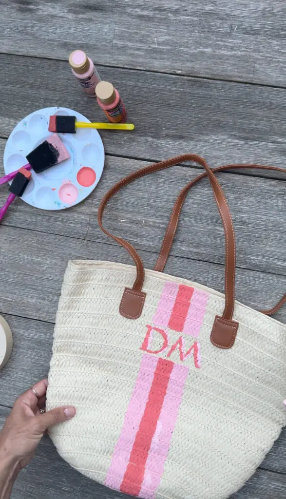 A hand holds a straw tote bag with brown handles, painted with pink stripes and the letters "DM." Nearby are paint supplies on a wooden surface.