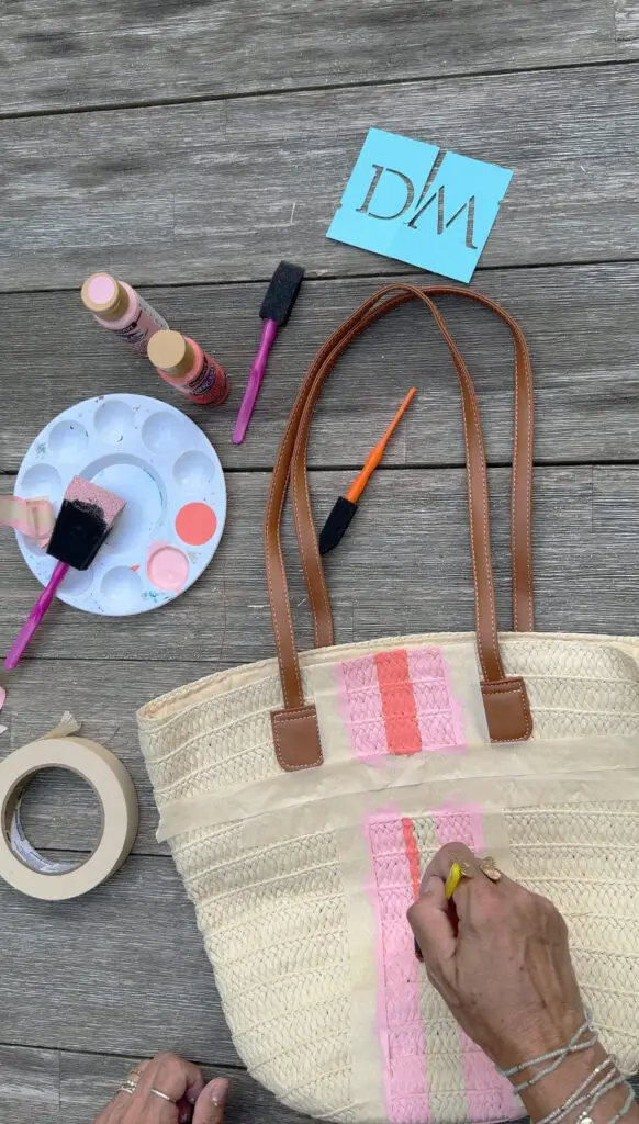A person paints pink stripes on a straw tote bag, with paint supplies, brushes, a stencil, and tape arranged on a wooden surface.