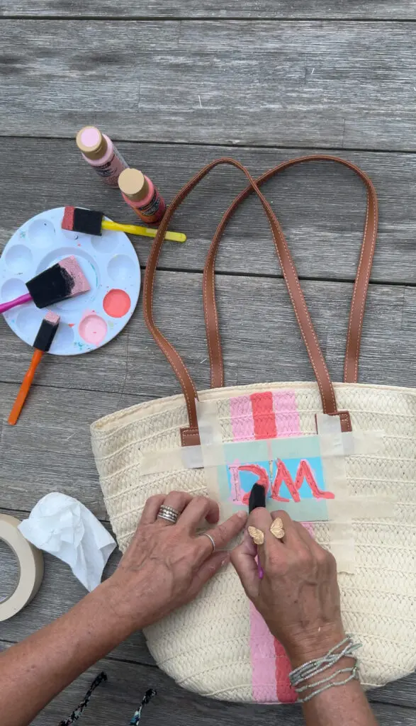 A person paints pink and blue letters on a woven straw bag with brown handles, using masking tape, paint, and brushes on a wooden surface.