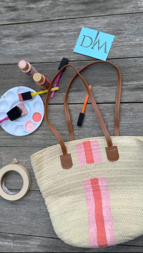 A woven tote bag with pink stripes is placed on a wooden surface, surrounded by paint supplies, a paint palette, tape, and a blue card with "DWA" printed on it.