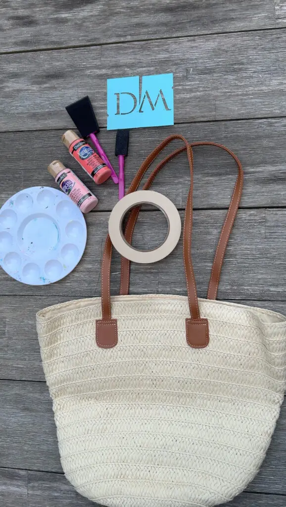 A straw tote bag, masking tape, paint palette, foam brushes, paint bottles, and a blue stencil with "DM" on a wooden surface.