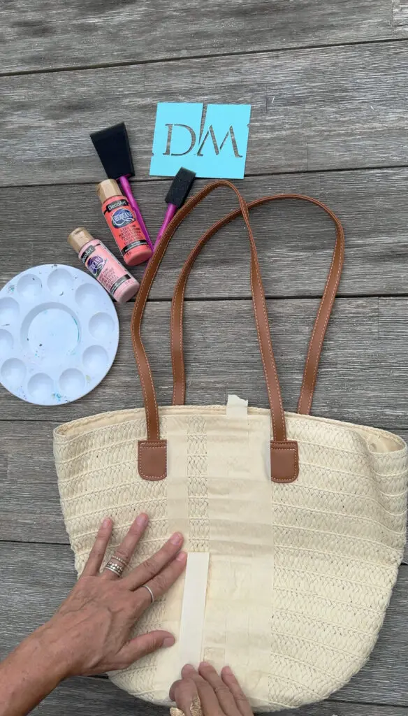 A straw tote bag with tan handles is taped off for painting; next to it are paintbrushes, acrylic paints, a palette, and a blue stencil reading "DM" on a wood surface.