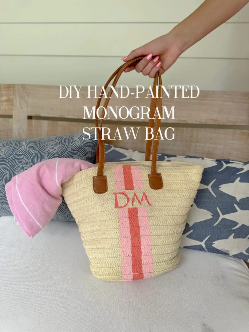 A hand holding a straw bag with brown handles, pink stripes, and "DM" initials sits on a bench next to a pink cloth and patterned pillows. Text reads "DIY Hand-Painted Monogram Straw Bag. Easy Paint Craft Project to Personalize Your Summer Bag with a DIY hand-painted monogram straw bag that is just so cute!